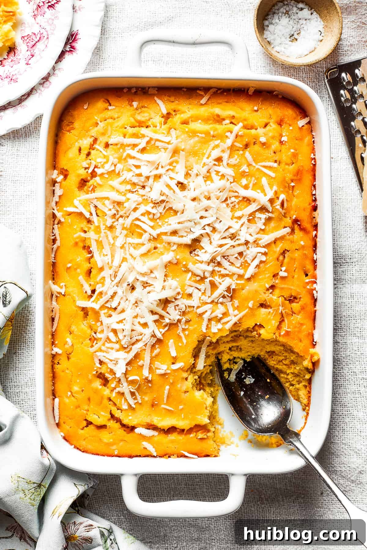 A white baking pan of baked corn souffle with a piece cut out and a spoon in the lower right side, showcasing its golden-brown top and creamy interior.