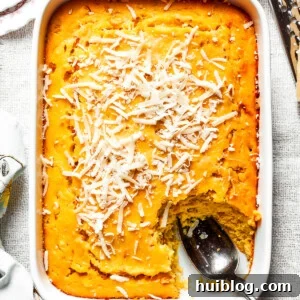 A white baking pan of baked corn souffle with a piece cut out and a spoon in the lower right side, showcasing its golden-brown top and creamy interior.
