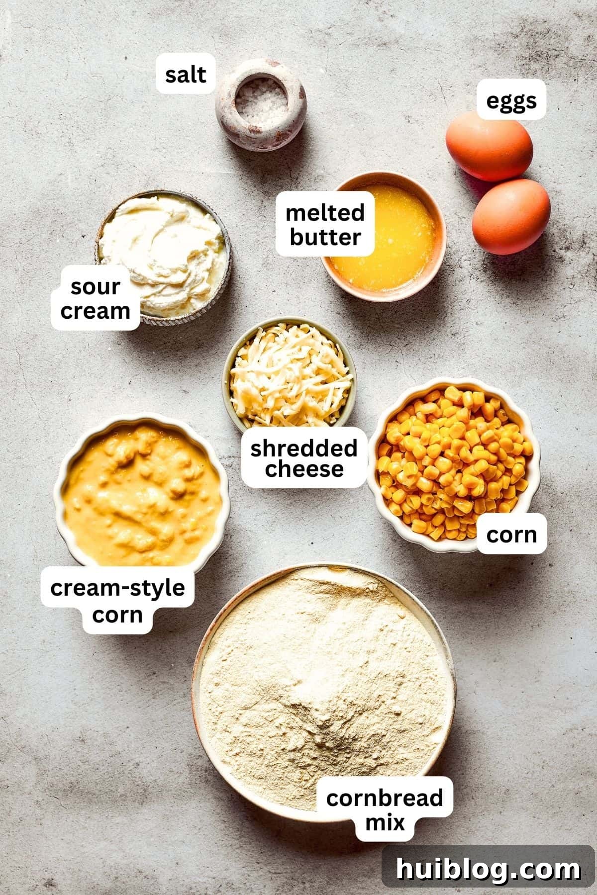 An overhead image of the ingredients used for Corn Souffle, including eggs, sour cream, butter, canned corn, cream-style corn, cornbread mix, and shredded cheddar cheese, neatly arranged on a white surface.