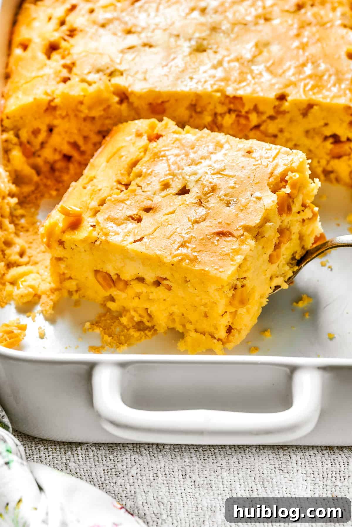 A perfectly cut-out square of golden-brown corn souffle, revealing its moist and creamy interior, resting within a white baking dish.