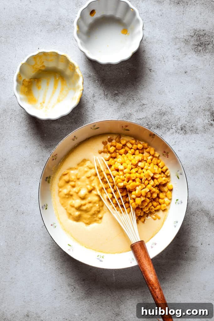 Whole kernel corn and creamed corn are added to a large mixing bowl containing the whisked souffle base ingredients.
