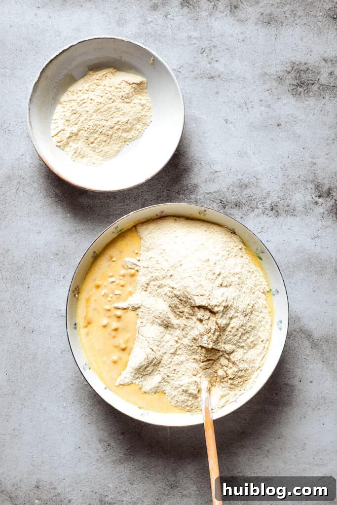 Cornbread mix is carefully poured into a bowl containing the prepared corn souffle base mixture, ready for stirring.