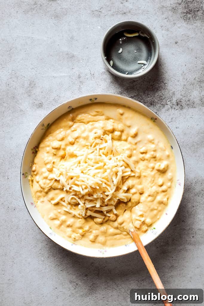 Shredded cheddar cheese is sprinkled into a bowl of corn souffle batter, adding a savory twist to the creamy mixture.