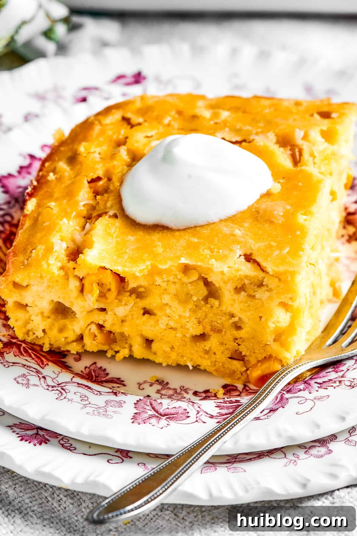 A square portion of golden-brown corn souffle served on a white dish, elegantly garnished with a dollop of sour cream and a sprinkle of fresh herbs.
