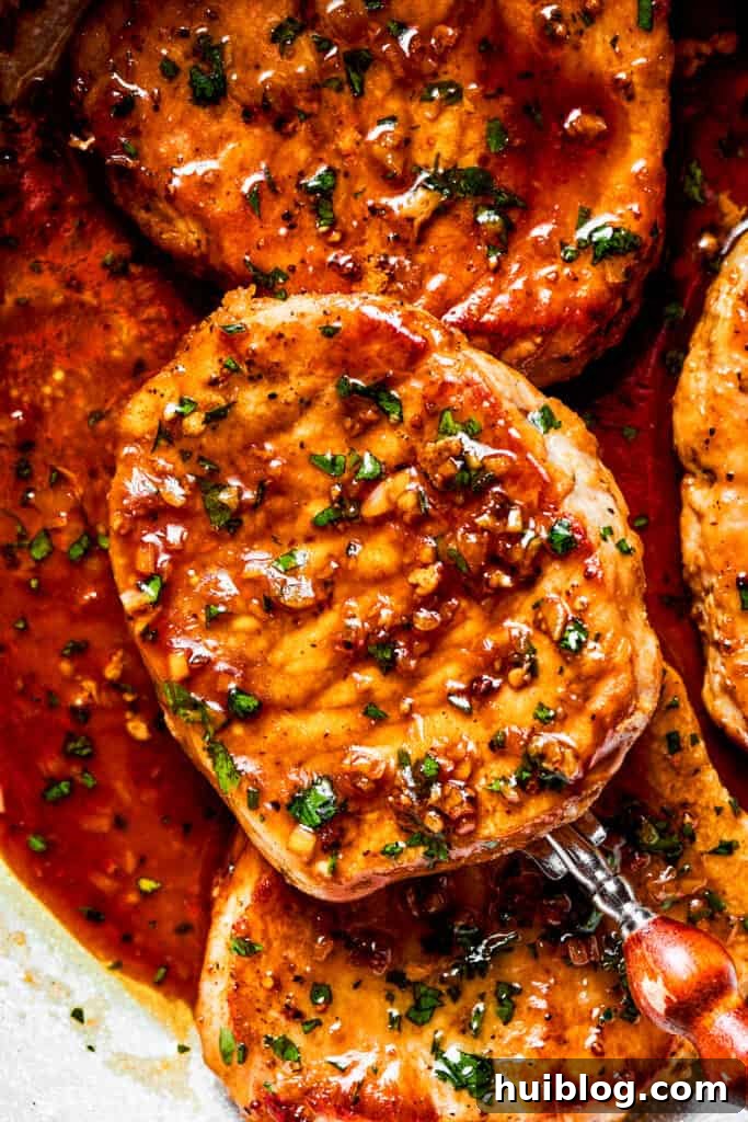 These Honey Garlic Pork Chops are the perfect blend of sweet-salty with a punch of extra flavor from minced garlic. Ready in 20 minutes! A close-up view of a wooden spoon lifting a perfectly seared honey garlic pork chop from the skillet, showcasing its beautiful glaze and tender texture.