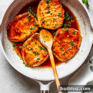 Four honey garlic pork chops cooking in a skillet with a wooden spoon stirring through.