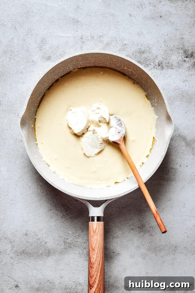 A wooden spoon stirs in cream cheese to a cream-butter mixture in a white skillet.