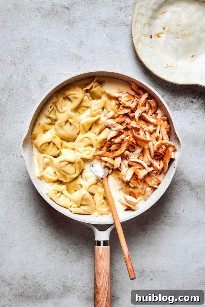 Cheese tortellini and shredded rotisserie chicken are stirred with a wooden spoon into a skillet of cream sauce.