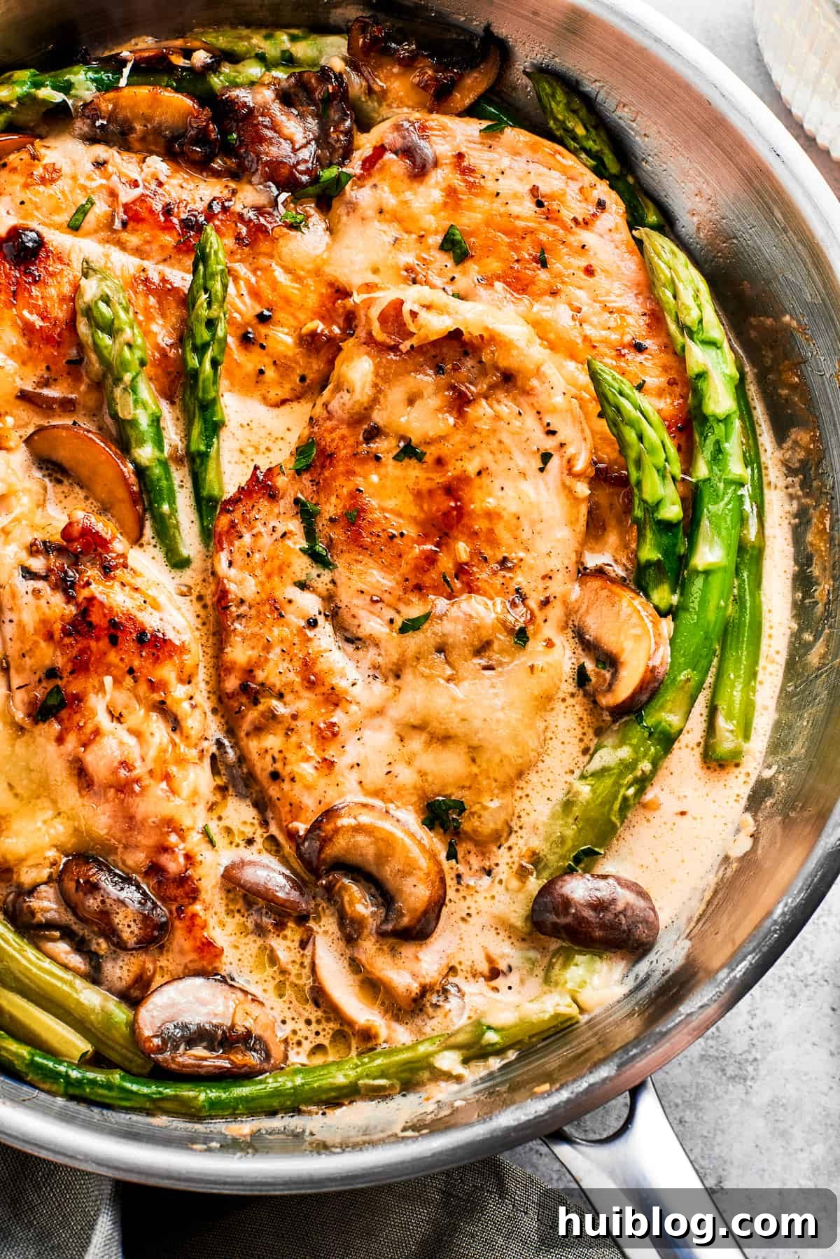 This creamy Chicken Madeira recipe is packed with savory sautéed mushrooms, sweet wine, tender chicken, and melted mozzarella cheese. Close-up photo of chicken Madeira and asparagus spears cooking in a skillet, showcasing the rich sauce and tender ingredients.