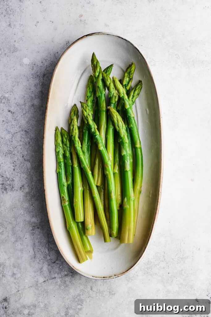 This creamy Chicken Madeira recipe is packed with savory sautéed mushrooms, sweet wine, tender chicken, and melted mozzarella cheese. Asparagus for chicken madeira are arranged in an oval white baking dish.