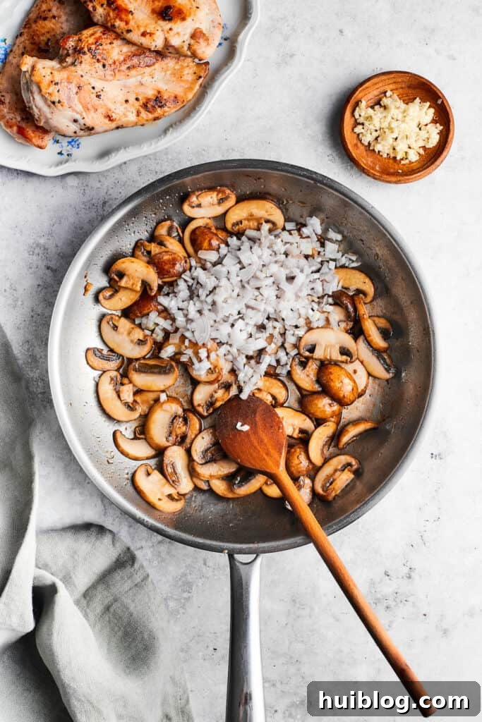 This creamy Chicken Madeira recipe is packed with savory sautéed mushrooms, sweet wine, tender chicken, and melted mozzarella cheese. Garlic is added to a skillet of cooking mushrooms with a wooden spoon.