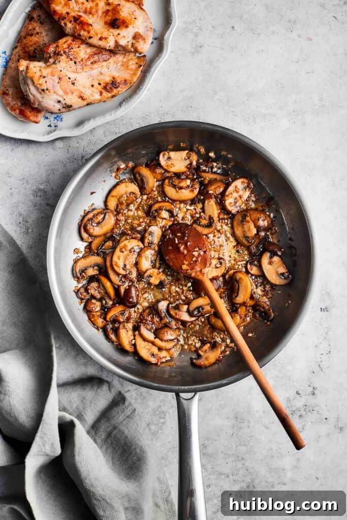 This creamy Chicken Madeira recipe is packed with savory sautéed mushrooms, sweet wine, tender chicken, and melted mozzarella cheese. A wooden spoon stirs a metal skillet full of cooked sliced mushrooms.