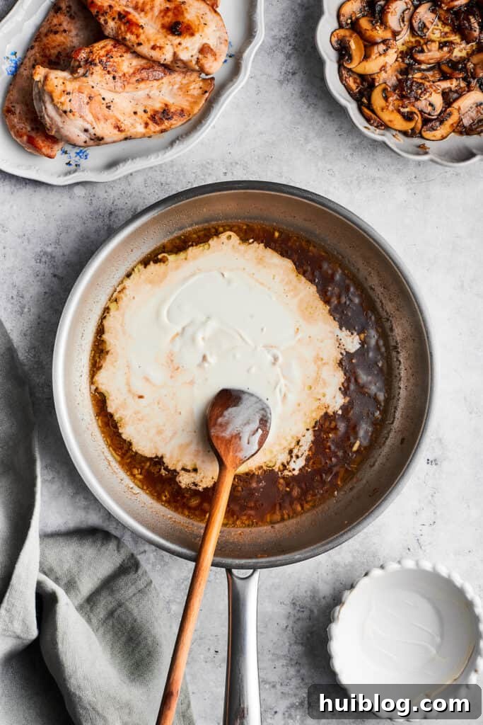 This creamy Chicken Madeira recipe is packed with savory sautéed mushrooms, sweet wine, tender chicken, and melted mozzarella cheese. A wooden spoon stirs in heavy cream into the sauce for chicken madeira.