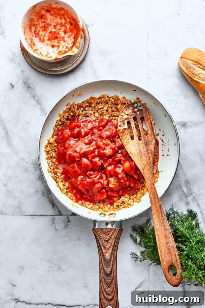 Thick diced tomatoes are stirred into a white skillet with a wooden spoon.