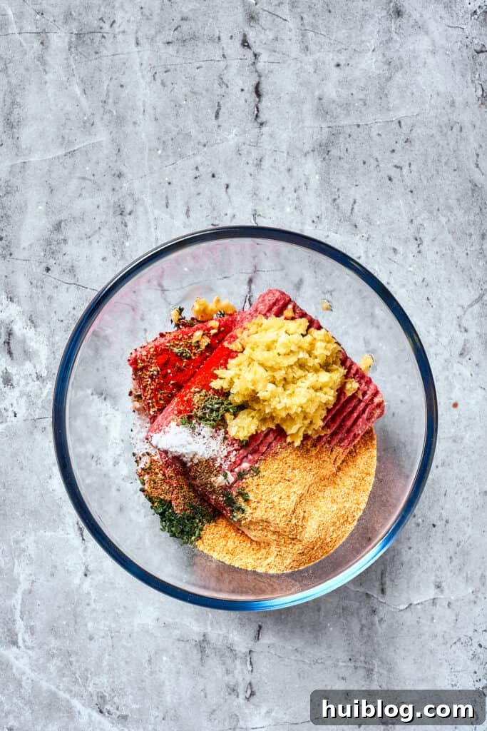 Comforting and hearty, Brown Sugar Meatloaf elevates ordinary meatloaf with a brown sugar-infused glaze for a hint of sweetness. A large clear glass bowl contains ground beef, mixed with breadcrumbs, cooked garlic and onions, and a medley of spices, ready for combining.