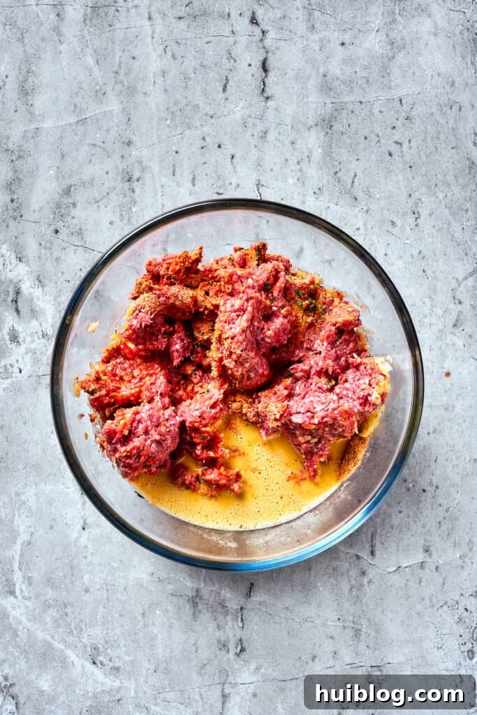 Comforting and hearty, Brown Sugar Meatloaf elevates ordinary meatloaf with a brown sugar-infused glaze for a hint of sweetness. The whisked egg mixture is gently poured into the large bowl containing the meatloaf base, ready to be incorporated by hand.