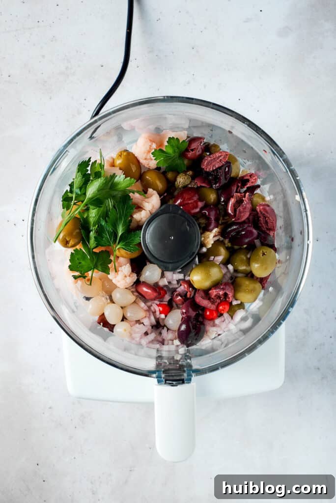 A mix of olives, peppers, pickled vegetables, and herbs being added to a food processor bowl.