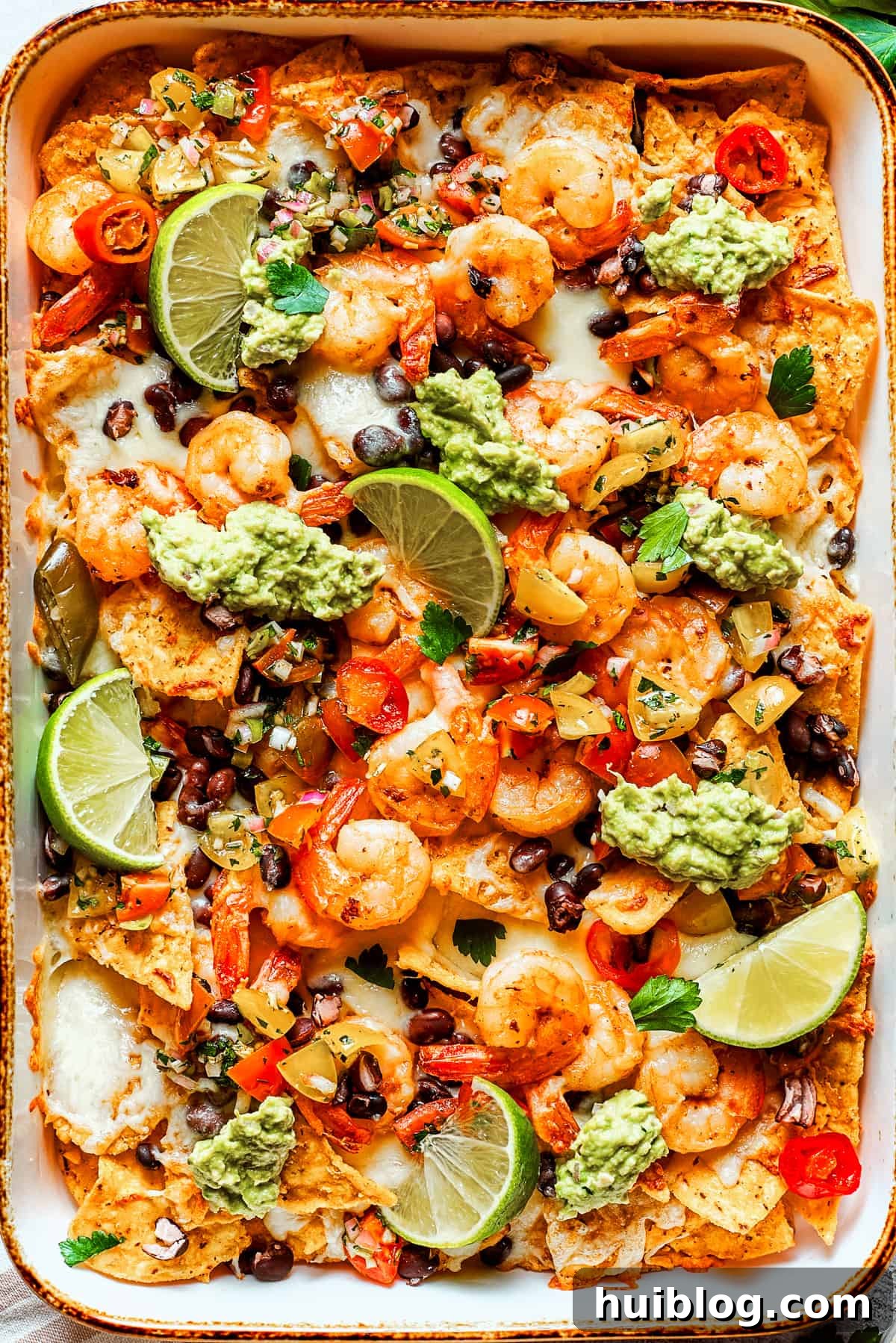 Coastal Crunch Shrimp Nachos 2 Nacho chips topped with shrimp, cheese, black beans, tomatoes, and a garnish of guacamole and slices of lime.