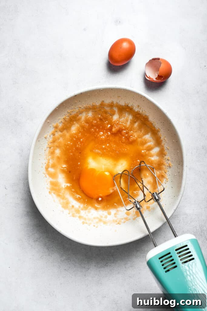 A hand mixer blending eggs into the oil and sugar mixture, ensuring a smooth emulsion.