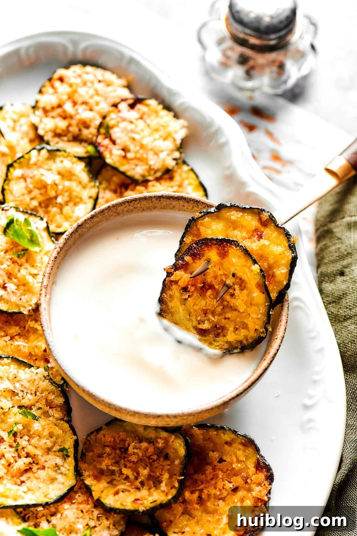 Two crispy zucchini chips resting in a small bowl of creamy yogurt dip, surrounded by more chips on a platter.