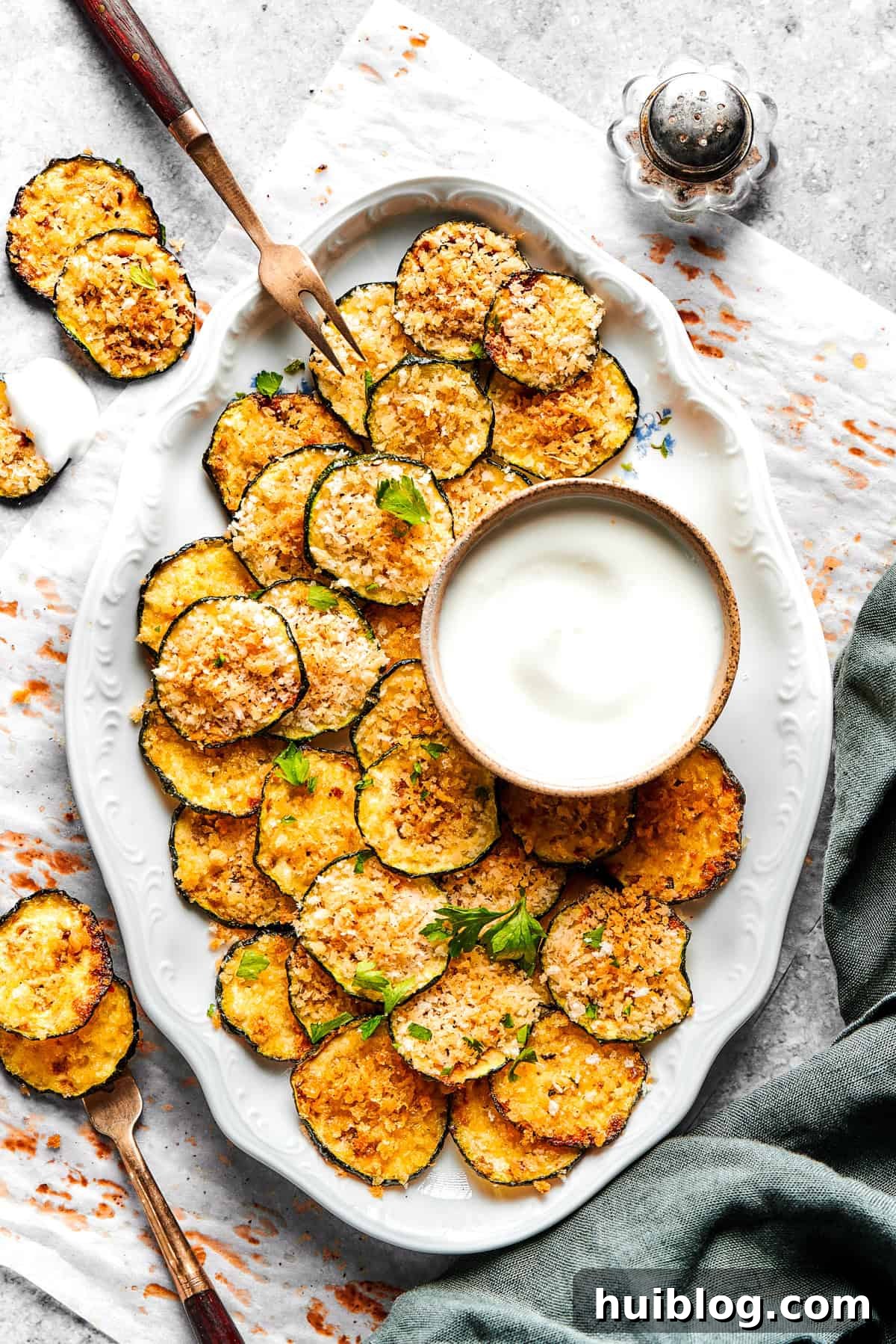 A beautiful serving of golden baked zucchini chips arranged on an oval white platter with a small bowl of creamy dip.