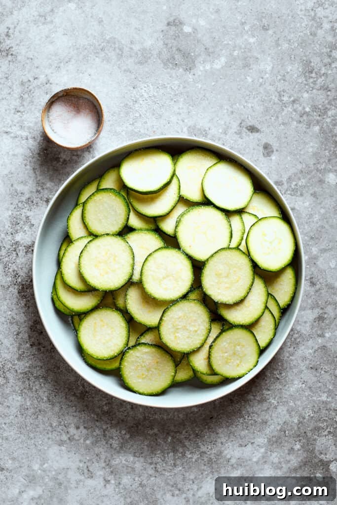 A bowl of uniformly sliced zucchini rounds next to a small bowl of sea salt, ready for preparation.