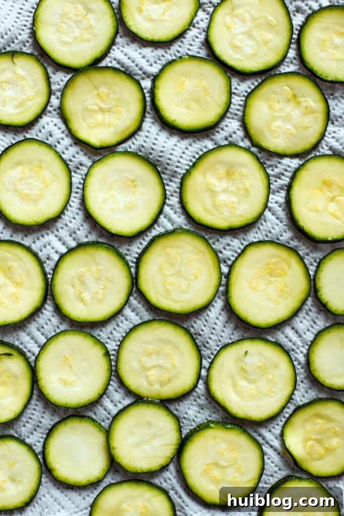 Dried zucchini slices are perfectly arranged in a single layer on a parchment-lined baking sheet, ready for oil.