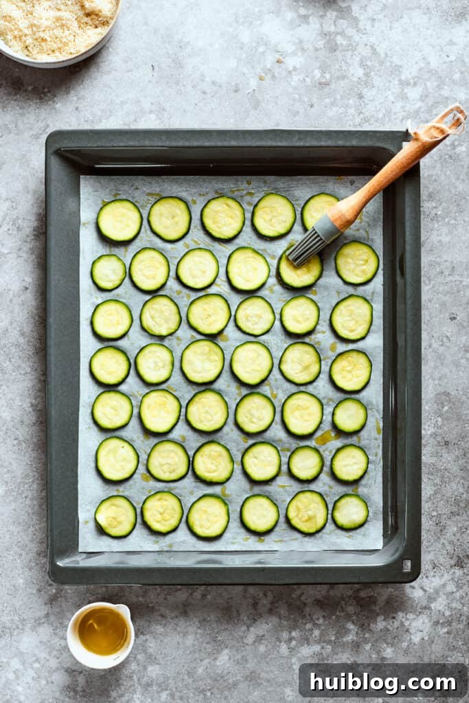 A pastry brush applies olive oil to zucchini chips on a baking sheet, ensuring an even coating.