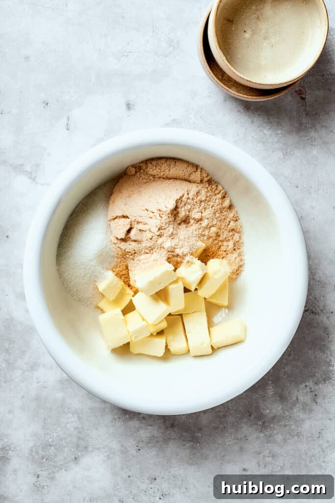 Butter, granulated sugar, and brown sugar are added to a white mixing bowl, ready to be creamed together.