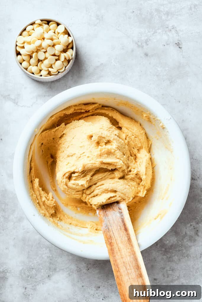 The white chocolate chip cookie dough is being gently stirred in a large mixing bowl, ensuring all ingredients are fully combined before adding the chocolate chips.