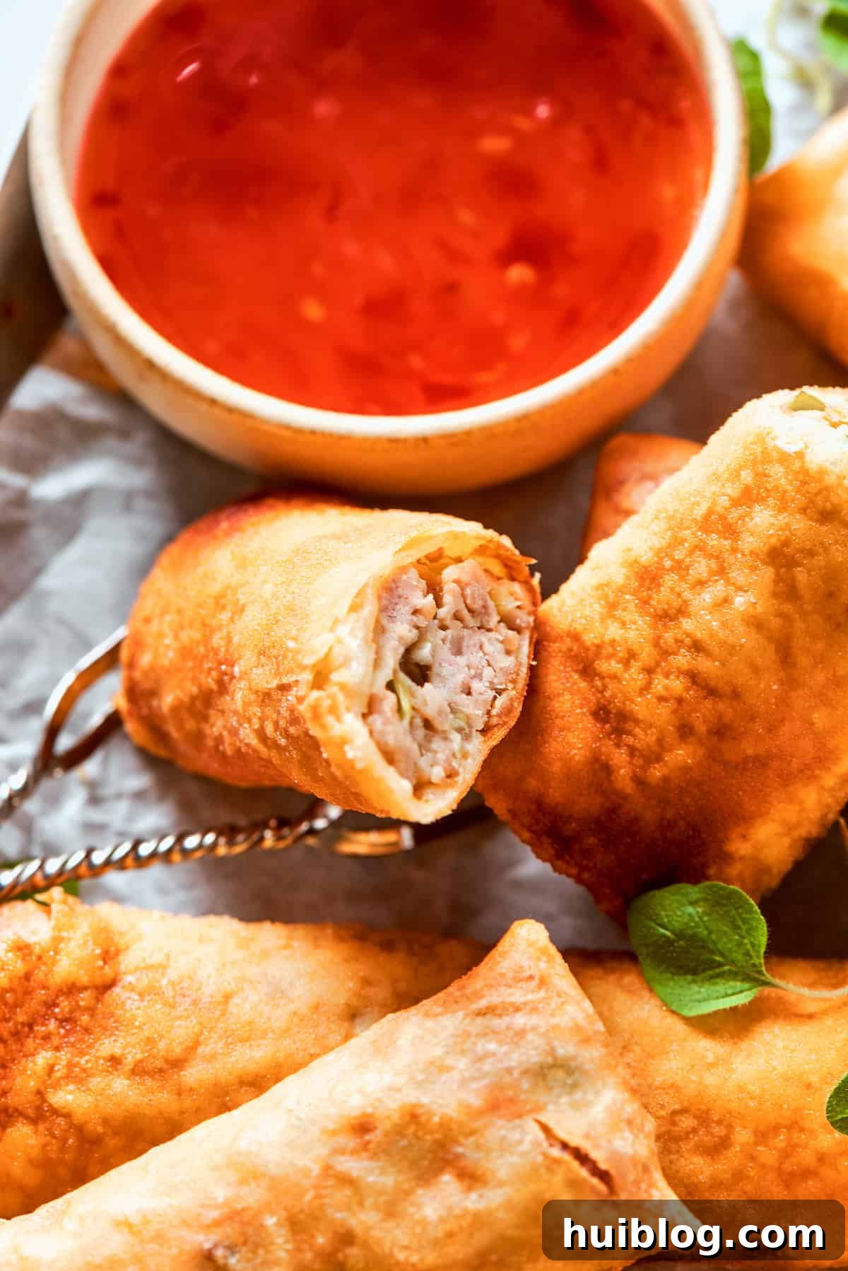 A pair of perfectly fried, golden-brown chicken egg rolls are presented on a white plate, with a small bowl of vibrant red sweet chili sauce positioned enticingly beside them, ready for dipping.