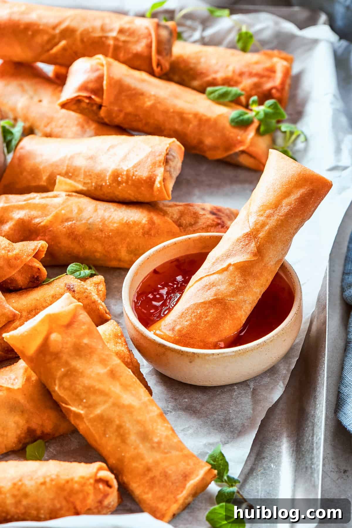 A crispy golden chicken egg roll is being dipped into a small, inviting bowl of red sweet chili sauce, with more delicious egg rolls scattered around on the serving surface.
