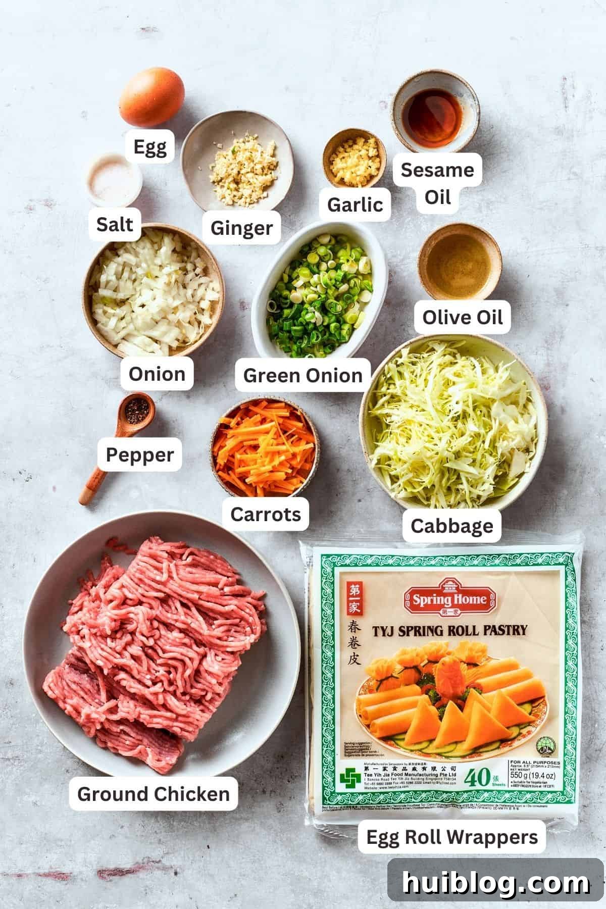 All the fresh ingredients for making crispy chicken egg rolls are neatly arranged and portioned out on a counter, including ground chicken, shredded carrots, cabbage, onion, garlic, olive oil, ginger, sesame oil, green onions, egg roll wrappers, salt, and pepper.