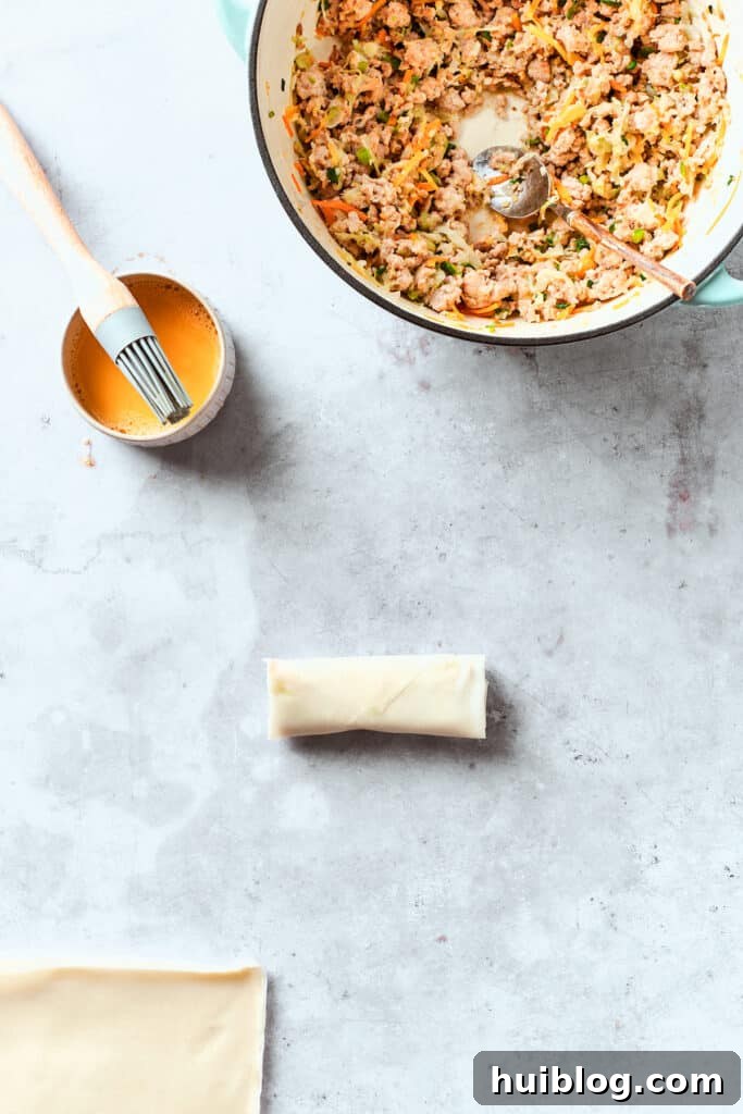 A light coating of beaten egg is being brushed onto the top corner of a neatly folded chicken egg roll, ensuring a strong seal and extra crispiness when fried.