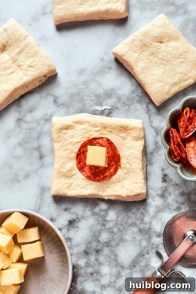 Carefully placing the filling onto each dough square. A slice of pepperoni and a cube of cheese is placed in the center of a dough square.
