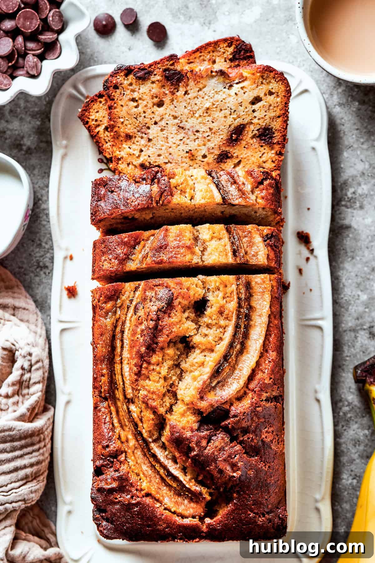 A perfectly baked loaf of Sour Cream Banana Bread, adorned with chocolate chips and a sliced banana on top, cooling in a loaf pan, surrounded by ripe bananas and other fresh ingredients.