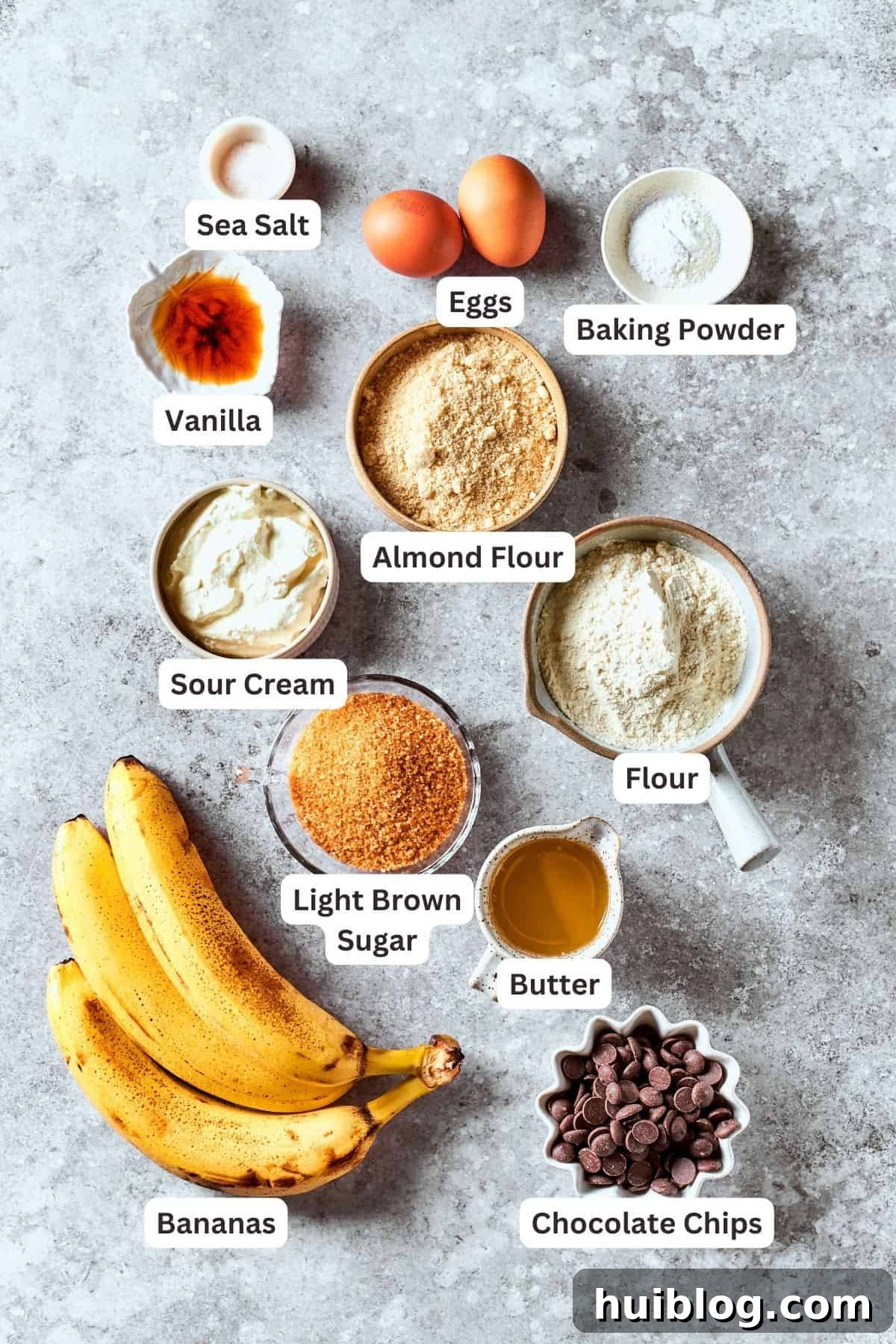 All ingredients for sour cream banana bread are neatly portioned and labeled on a kitchen counter: ripe bananas, brown sugar, almond flour, sour cream, all-purpose flour, baking powder, eggs, vanilla extract, salt, chocolate chips, and melted butter.