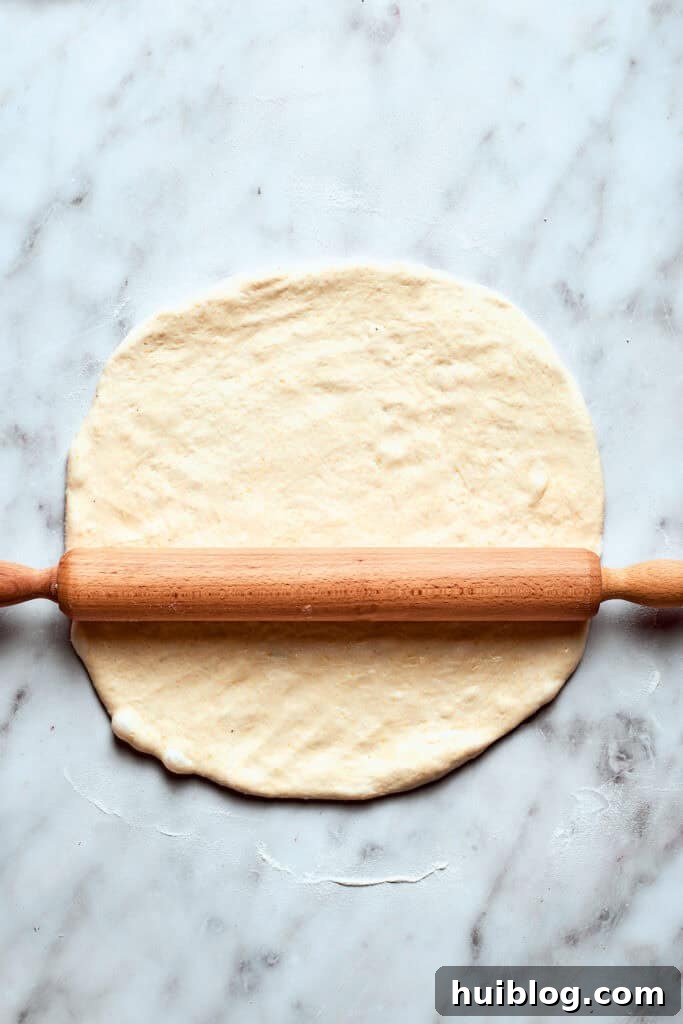 A rolling pin is used to roll out the risen deep dish pizza dough on a lightly floured surface.