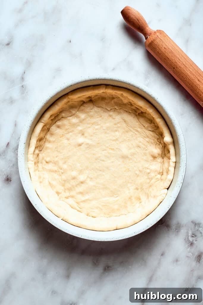 The rolled-out pizza dough is carefully pressed into a deep-sided baking pan, forming the base and sides of the deep dish.