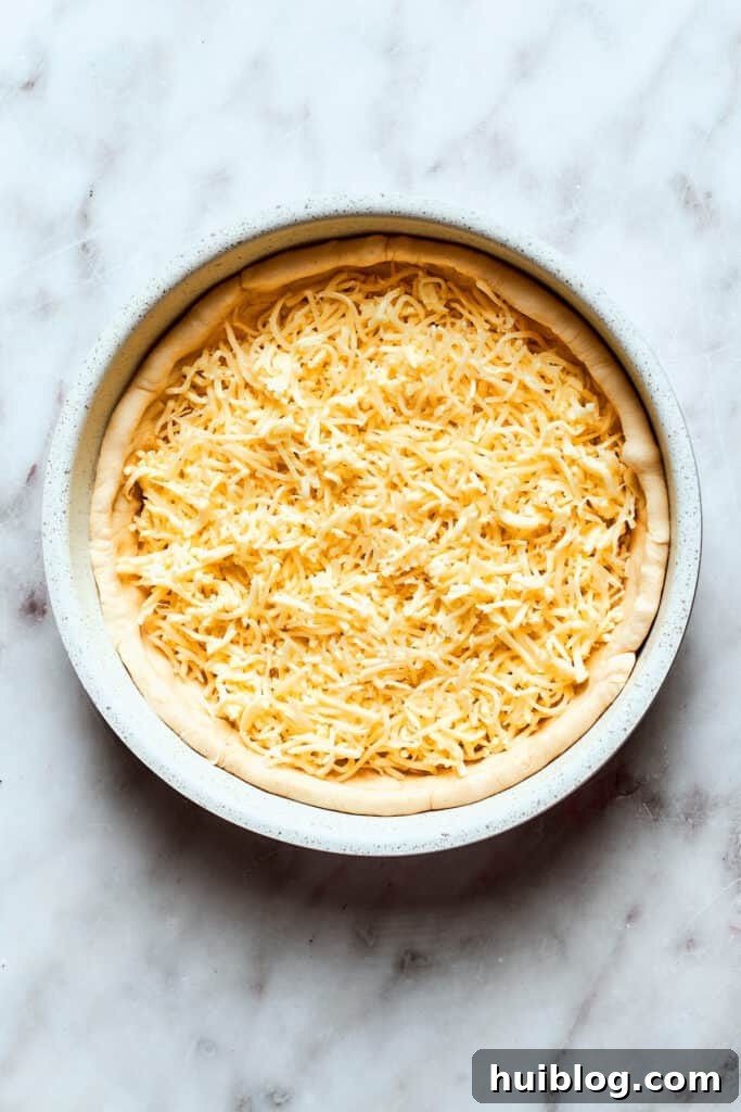 A generous layer of shredded cheese is spread evenly over the deep dish pizza dough, forming the first layer of toppings.