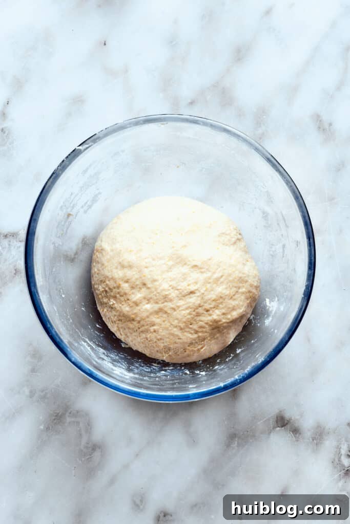 A perfectly kneaded pizza dough ball rests in a glass bowl, covered and rising in a warm place.
