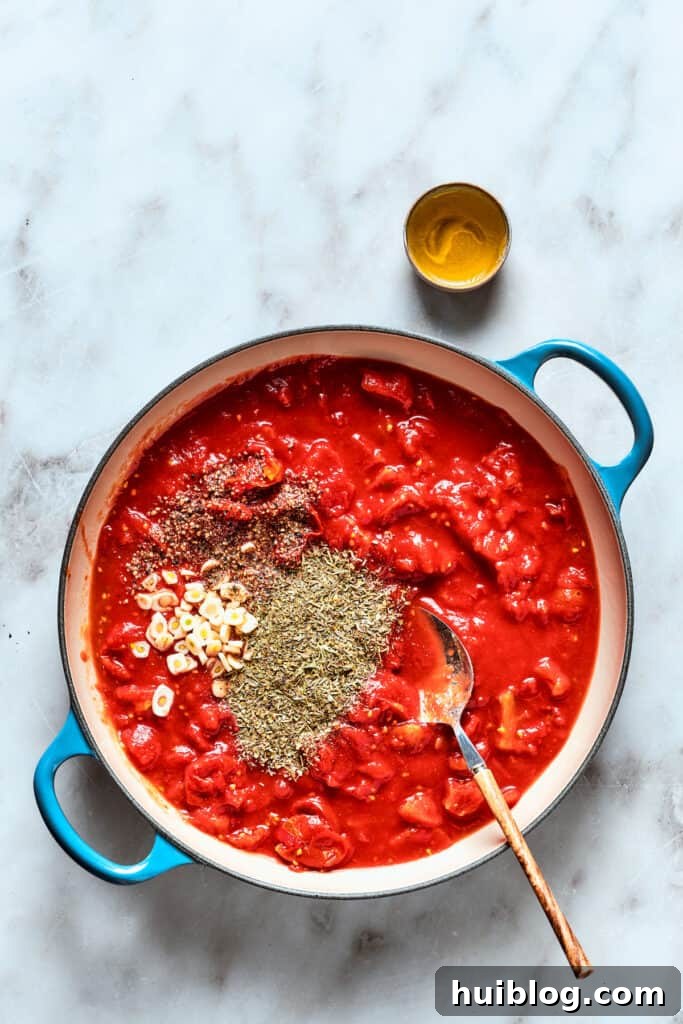 The rich, thick tomato sauce for deep dish pizza simmers in a saucepan, with Italian seasoning and other spices visible on the side, ready to be incorporated.