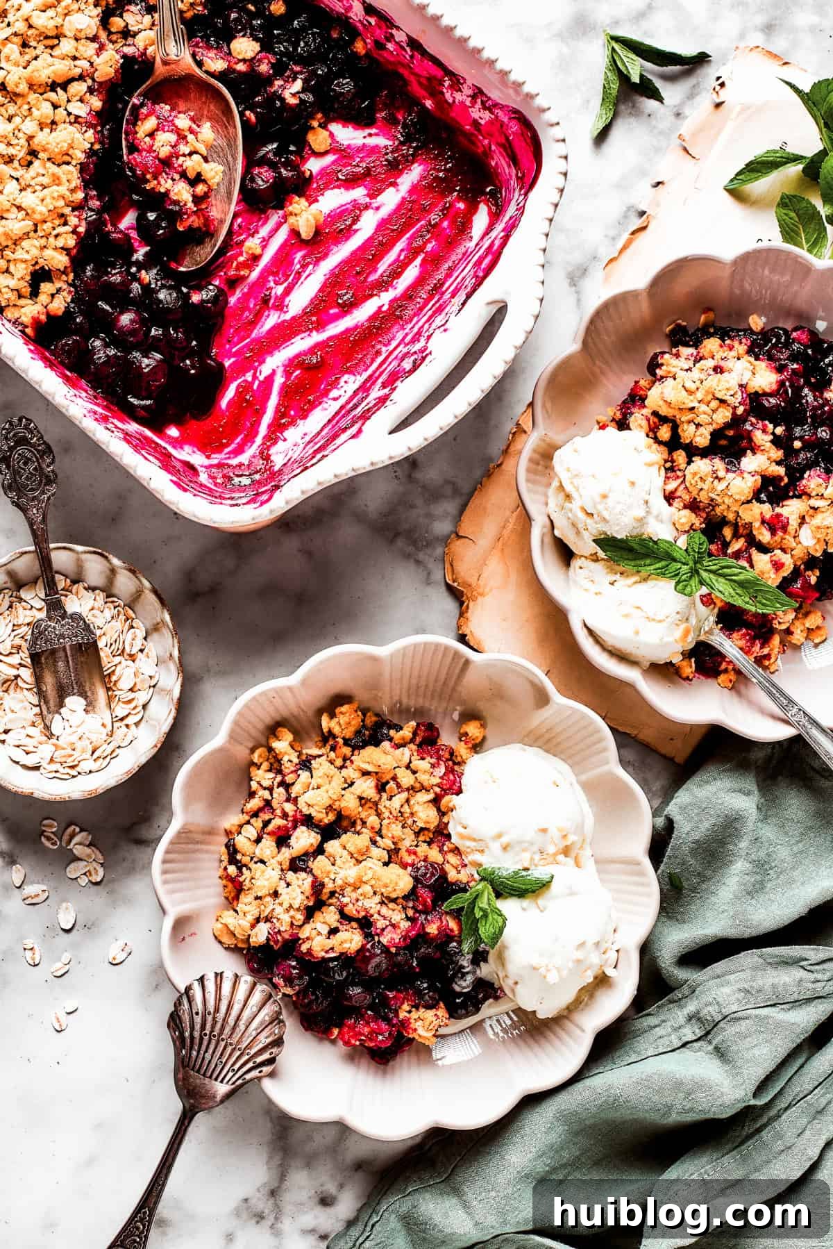 Overhead image of two dessert bowls of blueberry crisp served with scoops of ice cream. There is a baking dish with the crisp placed near the bowls.