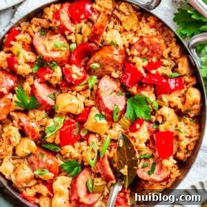 Image of a pan with jambalaya featuring a bed of rice topped with chicken bites, sliced sausages, and diced red bell peppers.