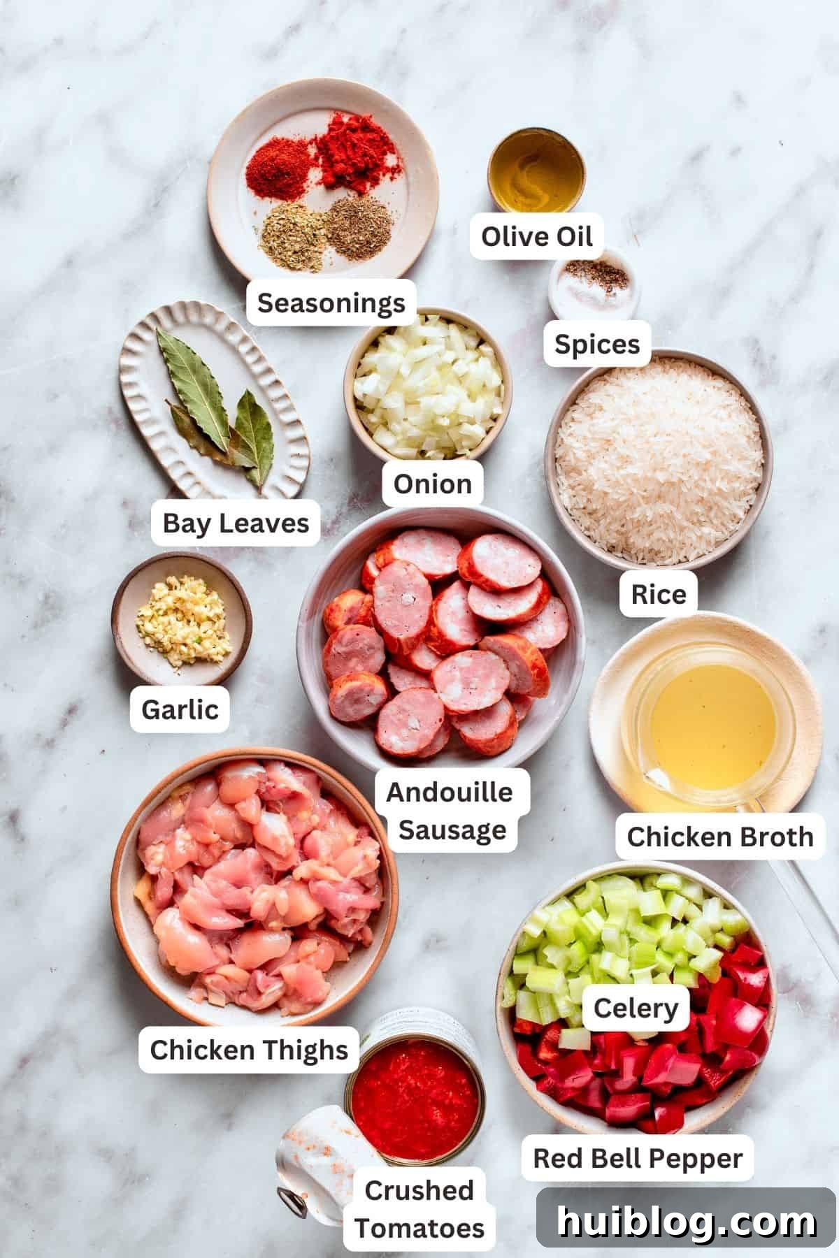 A beautifully arranged selection of fresh ingredients for Chicken and Sausage Jambalaya, including sliced Andouille sausage, raw chicken thighs, uncooked jasmine rice, garlic cloves, chicken broth, various seasonings and spices, olive oil, diced celery, and vibrant red bell pepper, ready for cooking.