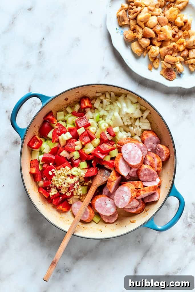 Diced onions, sliced sausage, red bell pepper, and celery are added to the pot after the chicken, beginning to sauté and release their aromas.