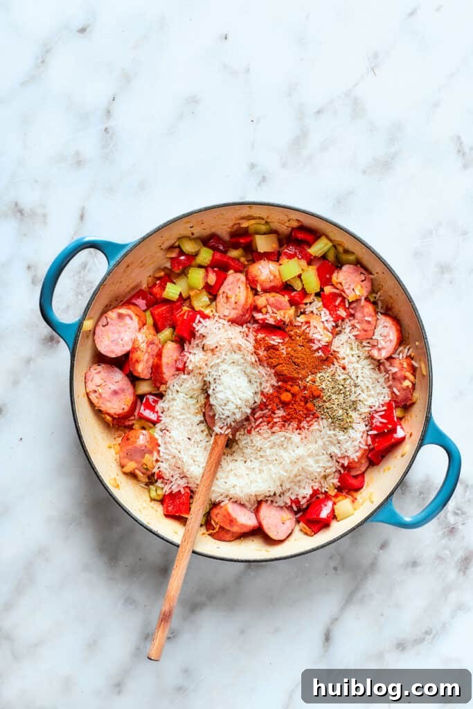 Uncooked rice and a blend of spices being sprinkled into the pot with sautéed vegetables and sausage, ready to be stirred.