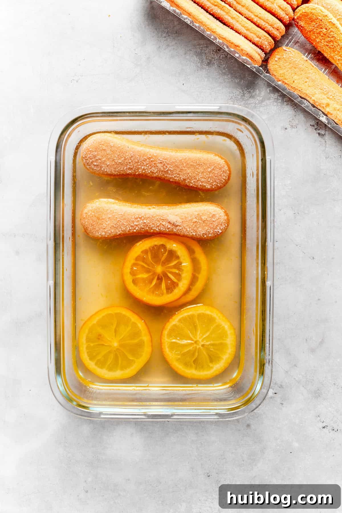 Ladyfinger cookies, lightly dipped in lemon syrup, are neatly arranged at the bottom of a baking dish, forming the first layer.