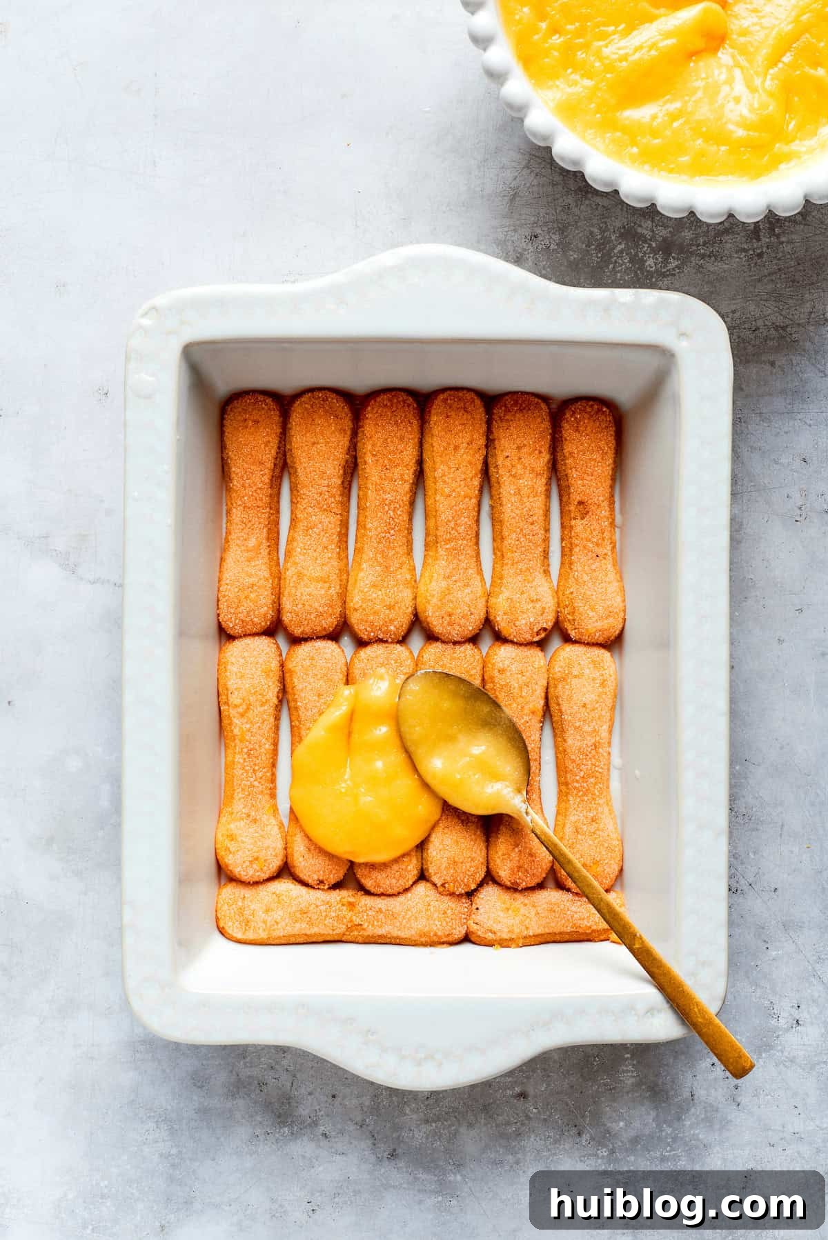 A smooth, even layer of homemade lemon curd being spread over the first layer of ladyfinger cookies in the baking dish.
