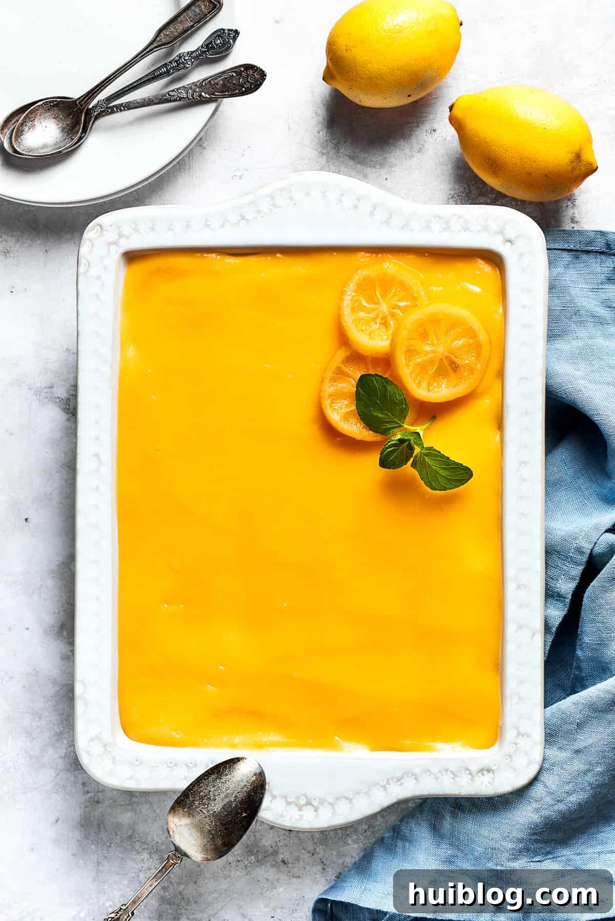 An overhead view of a finished lemon tiramisu in a baking dish, with fresh lemons arranged beside it for garnish.